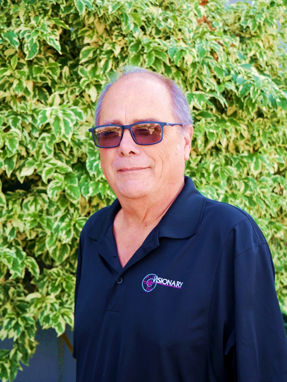 Man with glasses and grey hair standing in front of green bushes and a grey wall.